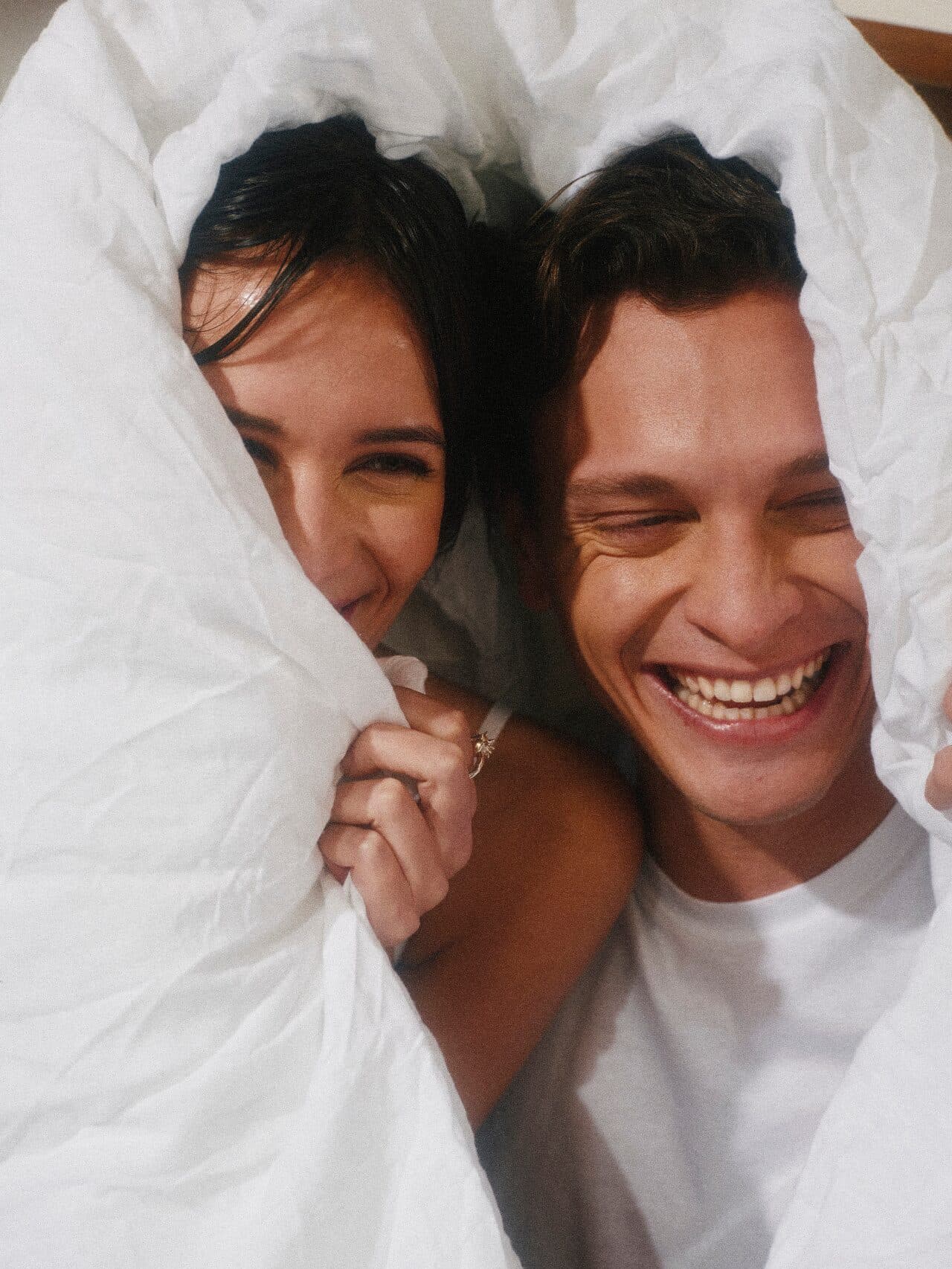 A couple laughing together beneath a white duvet