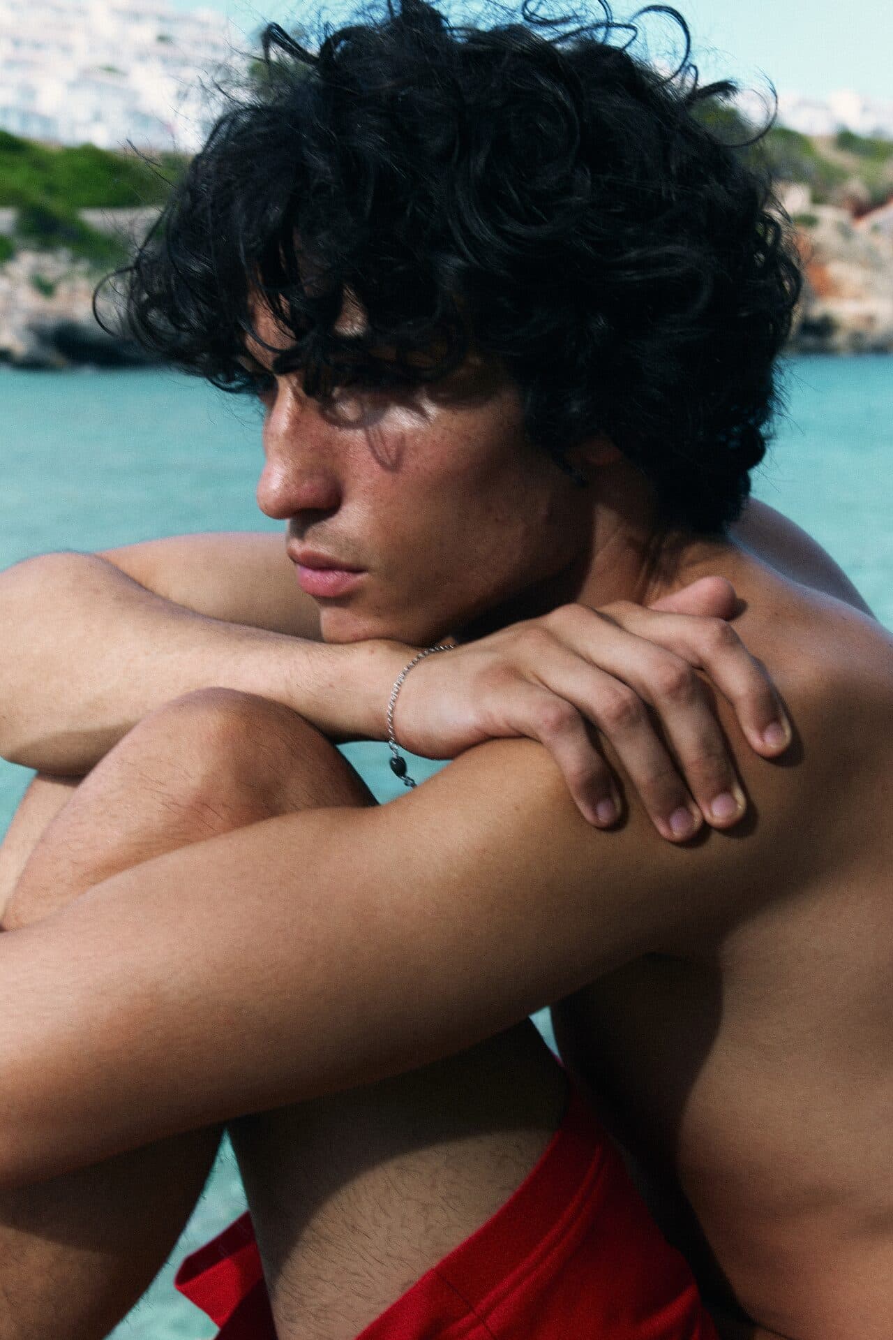 A man with thick curly hair sitting by the sea, arms resting on his knees