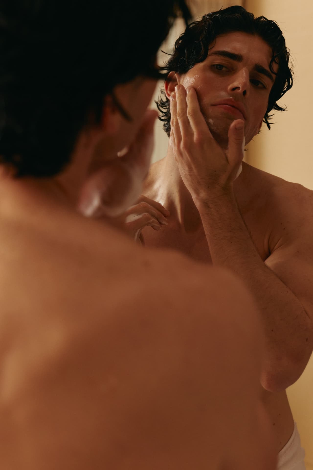A man studying his reflection in a bathroom mirror, hand resting on his jaw