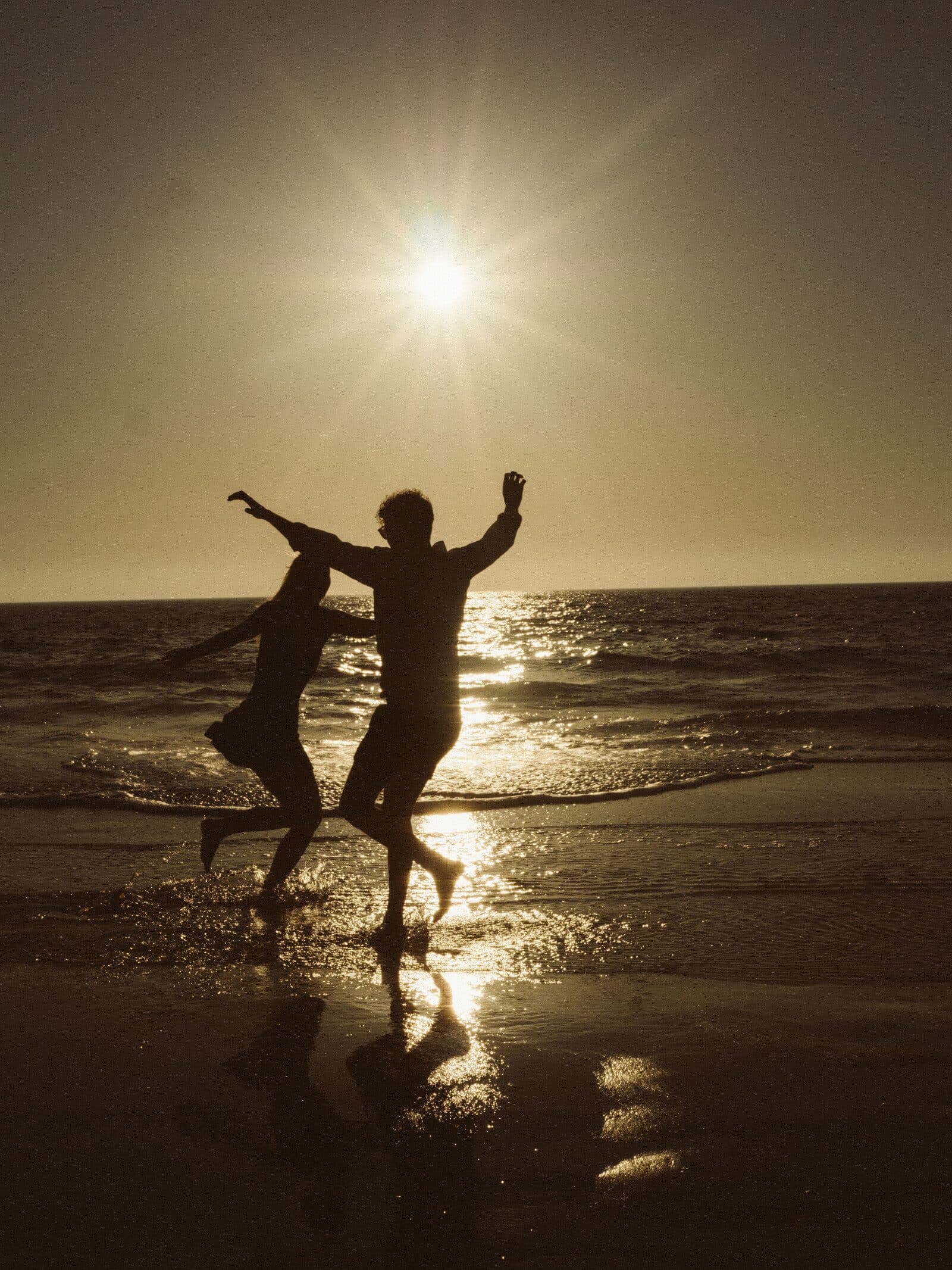 Two people leaping in the shallows, silhouetted against a low sun over the sea