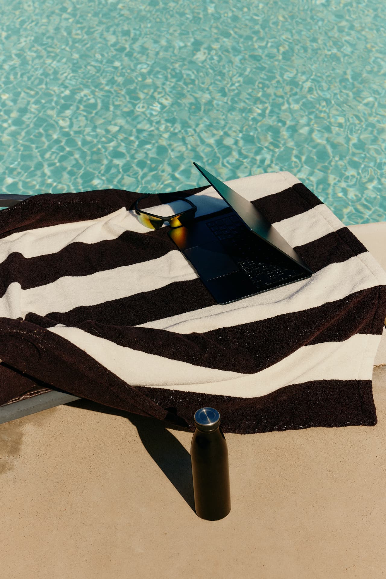 A striped towel and an open laptop beside a turquoise pool
