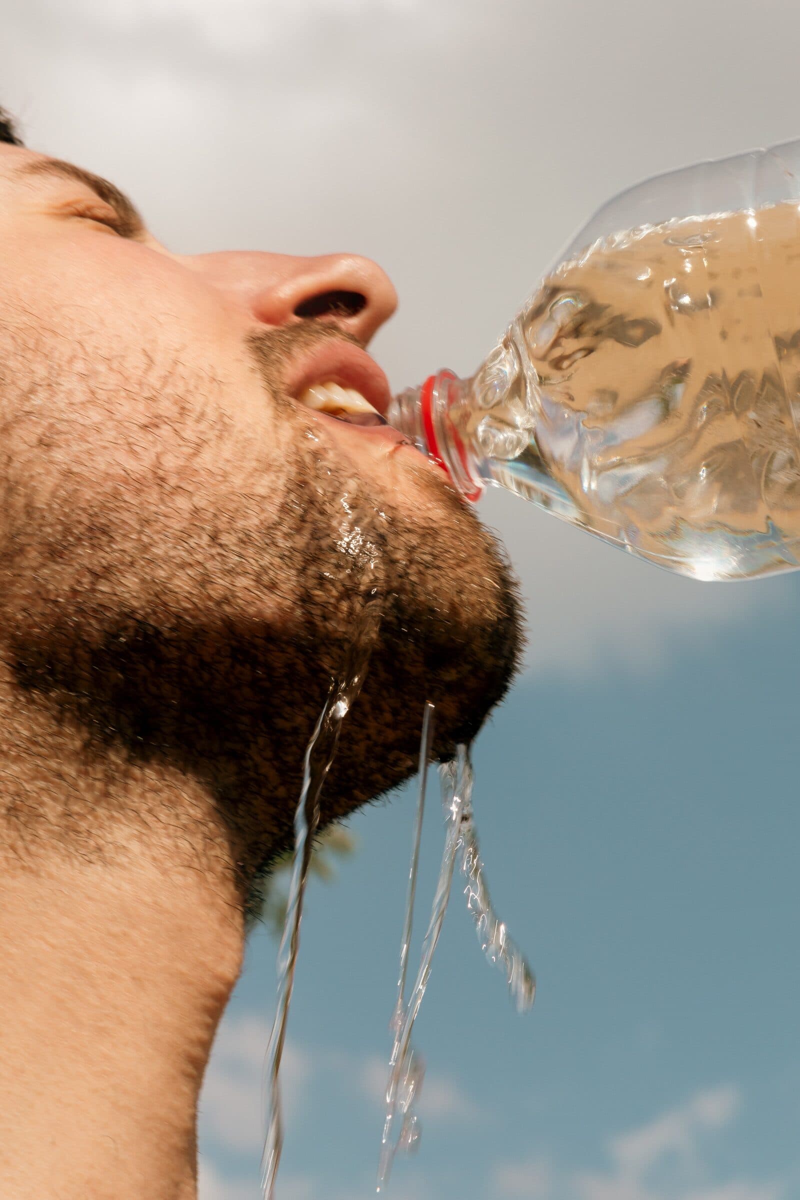 A bearded man tipping back a bottle of water against a bright blue sky